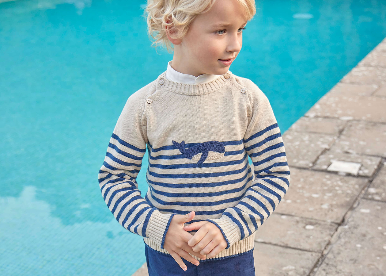 Young boy with blonde hair stands in front of a swimming pool. He is looking off to the side and his hands are clasped in front of him. He is wearing the Pepa London Whale Intarsia Striped Jumper and a pair of navy blue shorts.